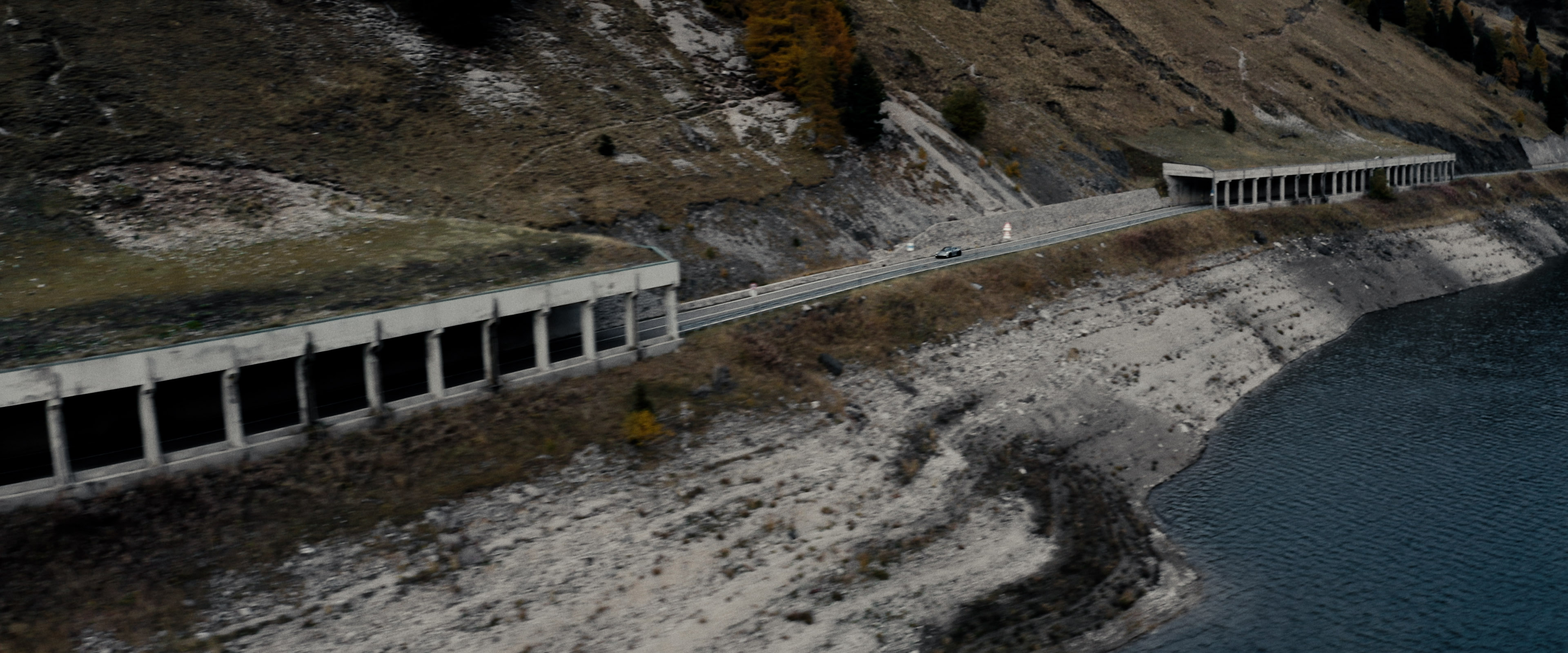 Aerial view of an alpine lakeside road with avalanche gallery; tiny sports car speeds along the shoreline.