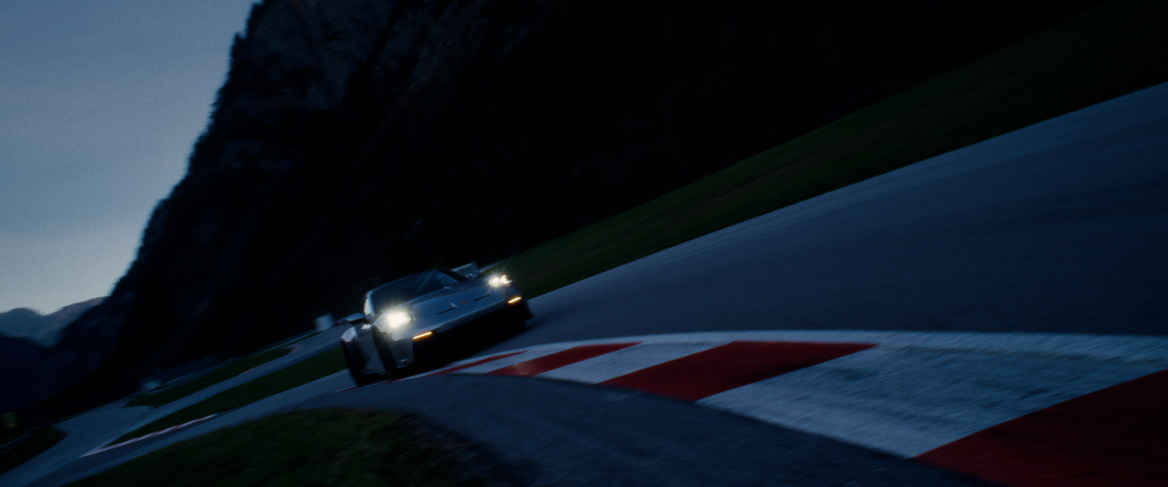 Front-facing Porsche GT3 with headlights on, carving through a chicane at twilight in alpine scenery.